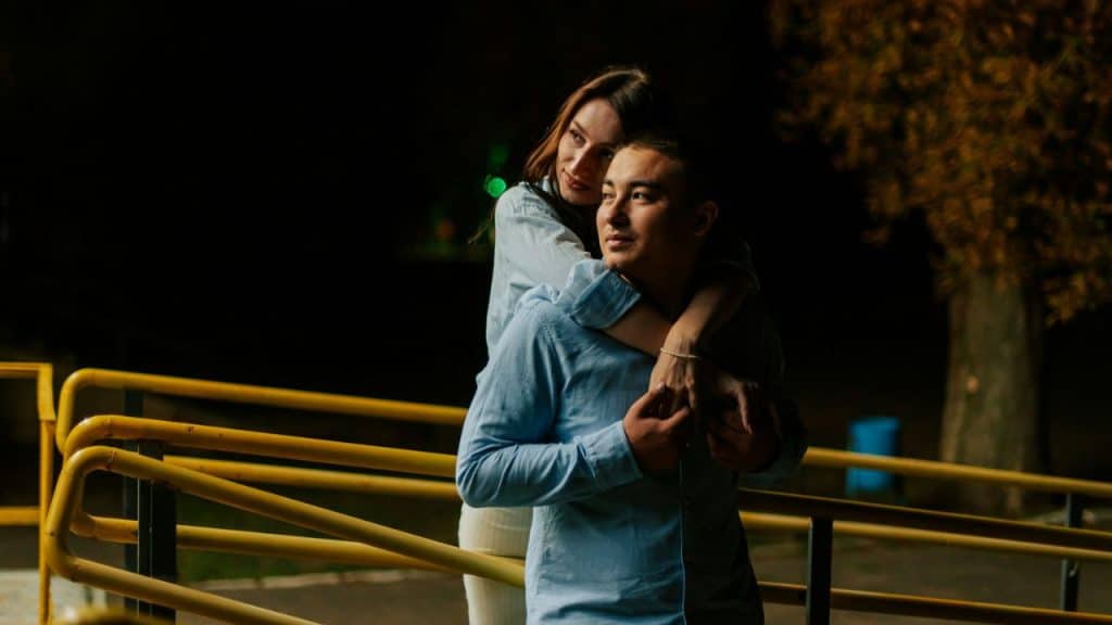 A woman in a blue shirt hugs a man in a blue shirt from behind outdoors.