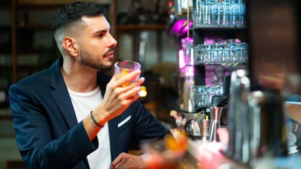 A bearded man in a navy blazer holds a drink at a brightly lit bar.