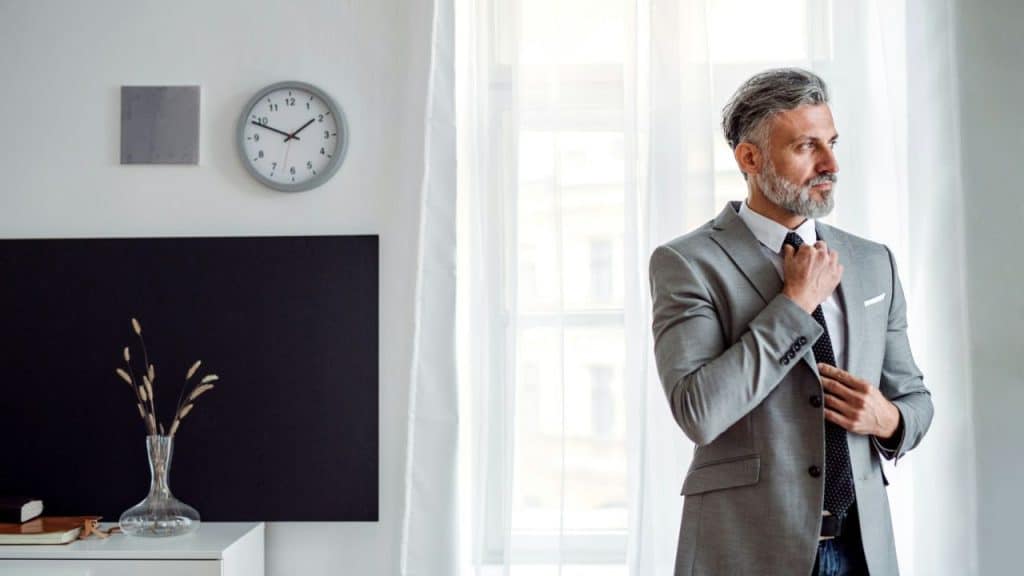 A gray-haired man in a light gray suit adjusts his tie while looking out a window.