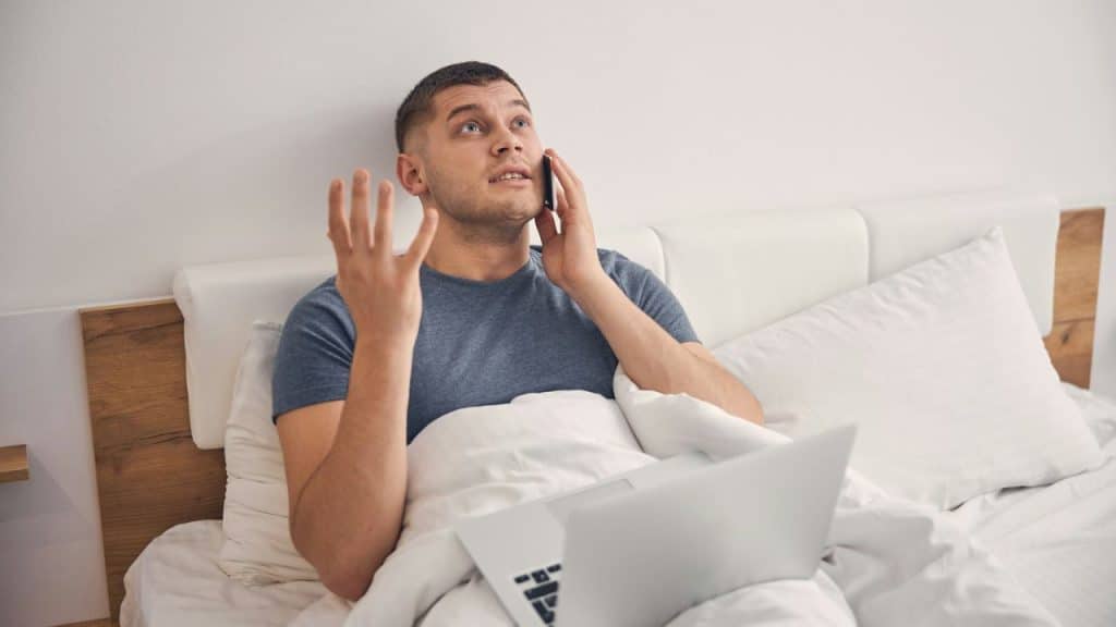 A man in a blue t-shirt sits in bed with a laptop, talking on a mobile phone.