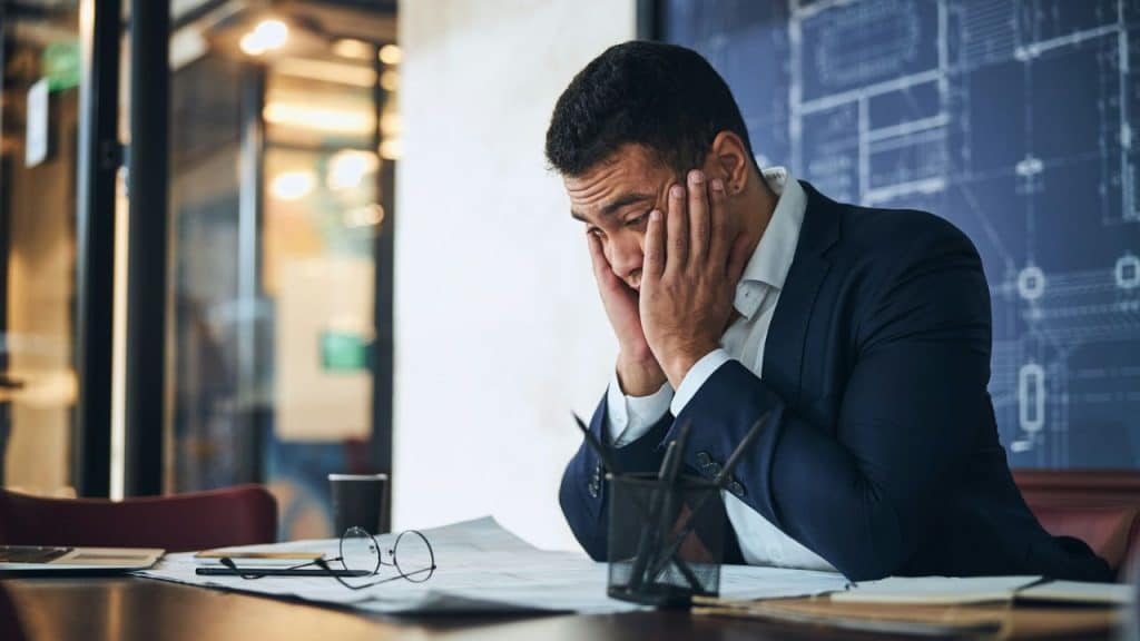 A man in a suit sits at a desk, holding his face in his hands over papers.