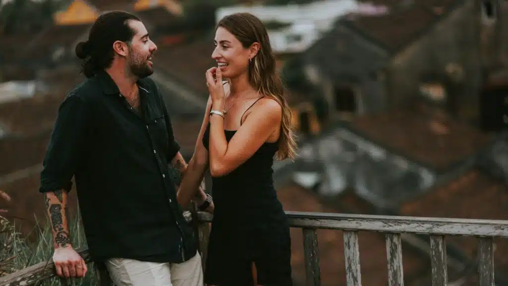 A man with a bun and a woman in a black dress laugh on a balcony.