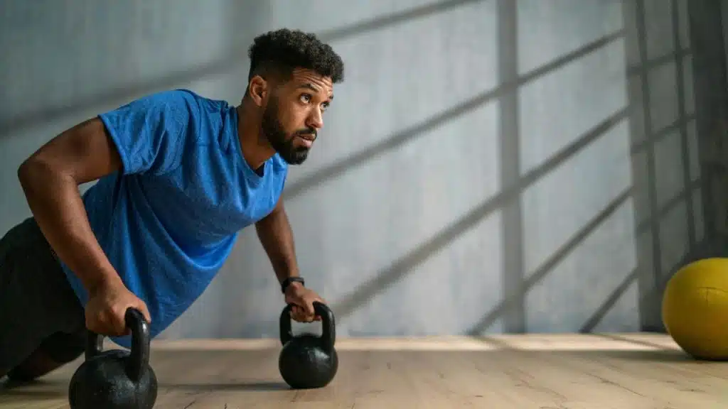 A man in a blue shirt performs push-ups using kettlebells in a sunlit gym.
