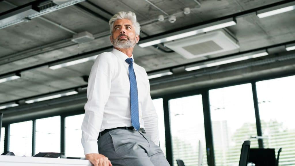 A man with silver hair and a beard looks upward while leaning against a desk.