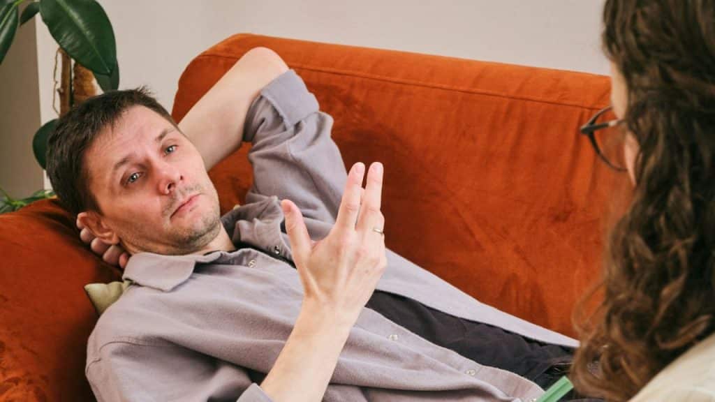 A man reclines on an orange sofa, gesturing with one hand while looking at a woman.