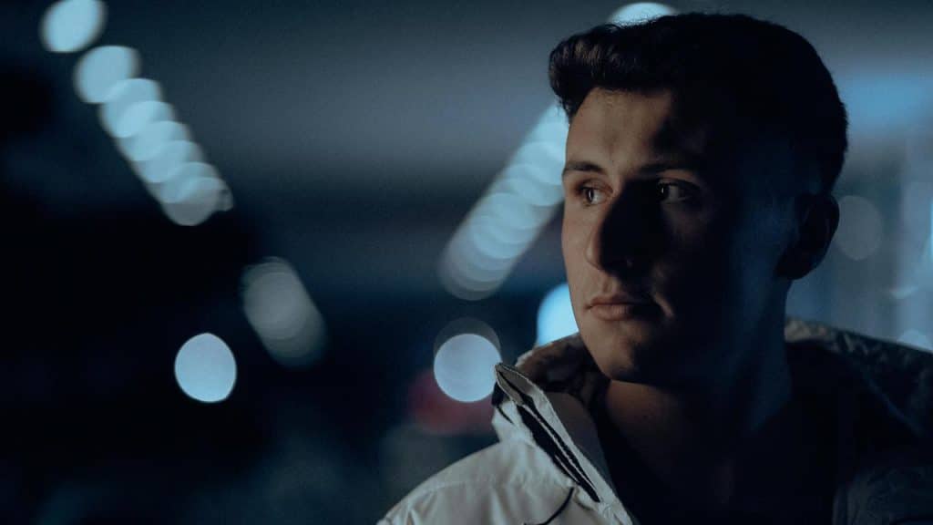 A man in a dimly lit space, looking to the side with blurred lights in the background.