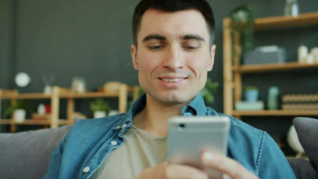 A man in a denim shirt smiles while looking down at a smartphone in his hands.
