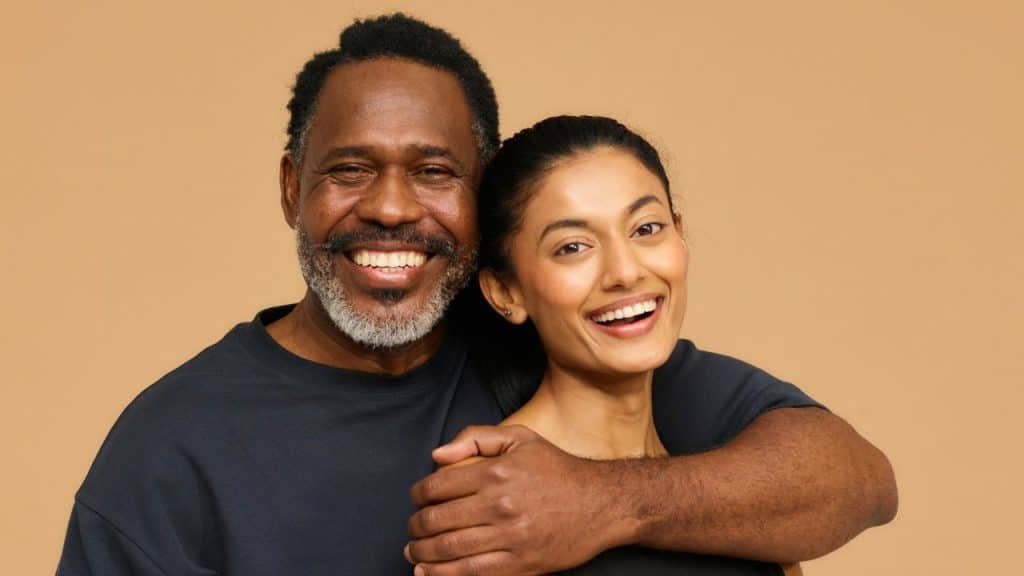 A smiling man with a salt-and-pepper beard embraces a smiling woman against a tan background.