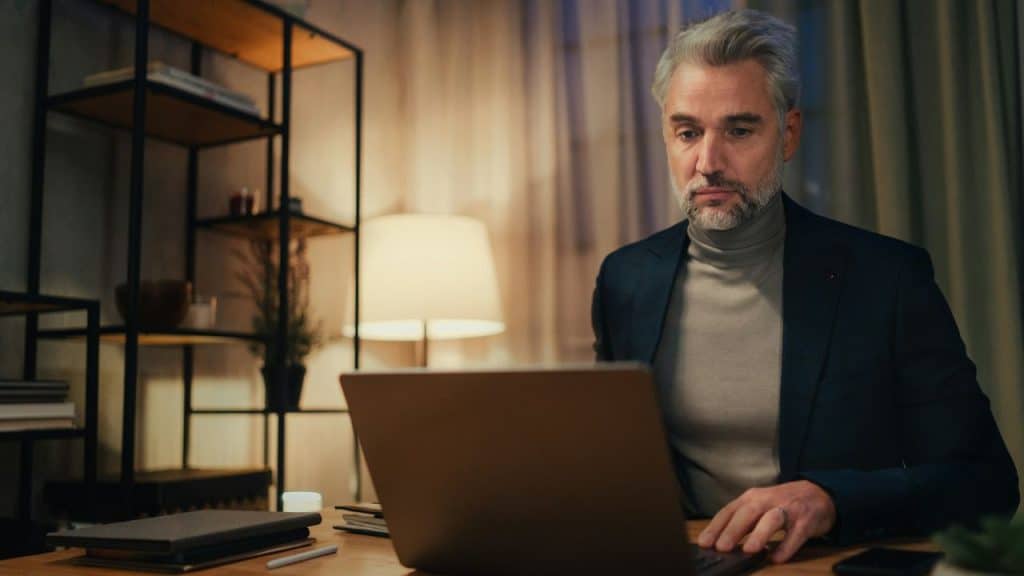 A man with gray hair sits at a desk working on a laptop in a dimly lit room.