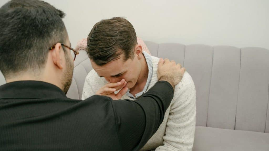 A person in a black shirt comforts another person who is crying on a couch.