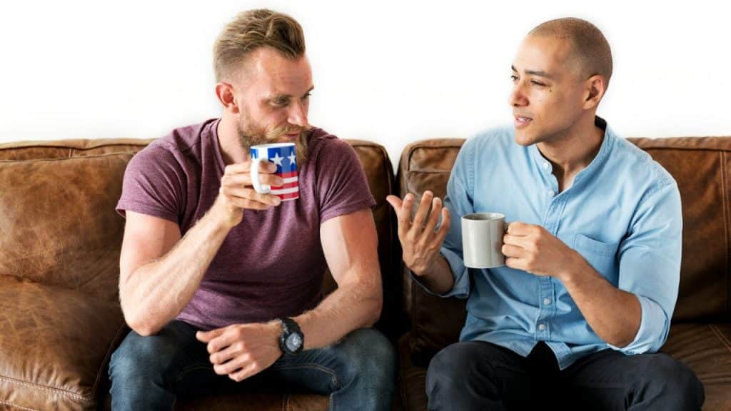 Two men sit on a leather couch, holding mugs and engaging in a conversation together.