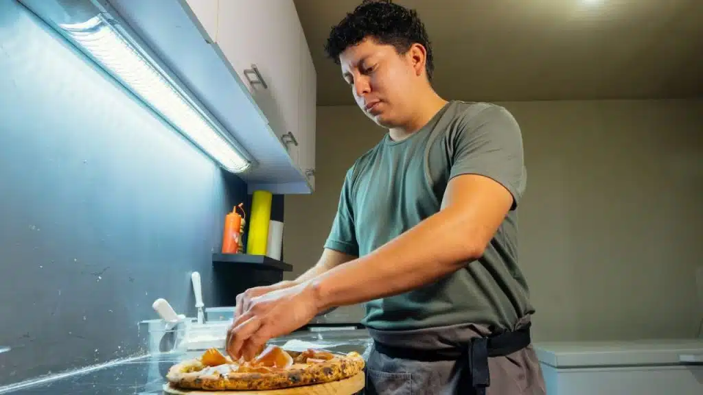 A man in a green shirt prepares food on a circular crust in a kitchen.