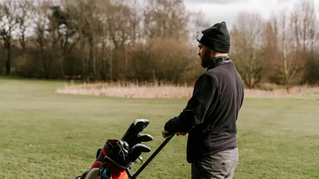 A man in a beanie pushing a golf bag across a grassy course on a cloudy day.