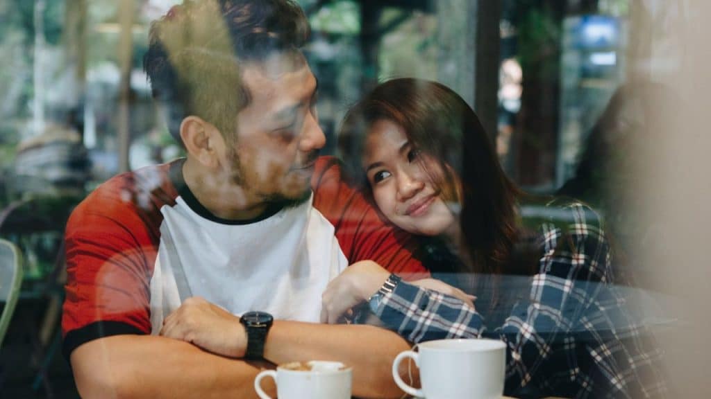 A man and woman lean against each other at a table, looking through a window.