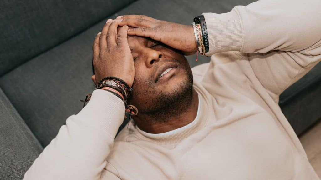 A man lies back with eyes closed, pressing his hands against his forehead and temples.