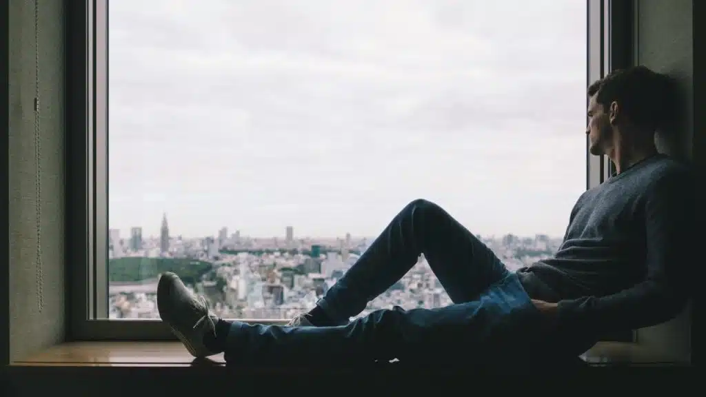 A man sitting on a window ledge looking out at a cityscape.