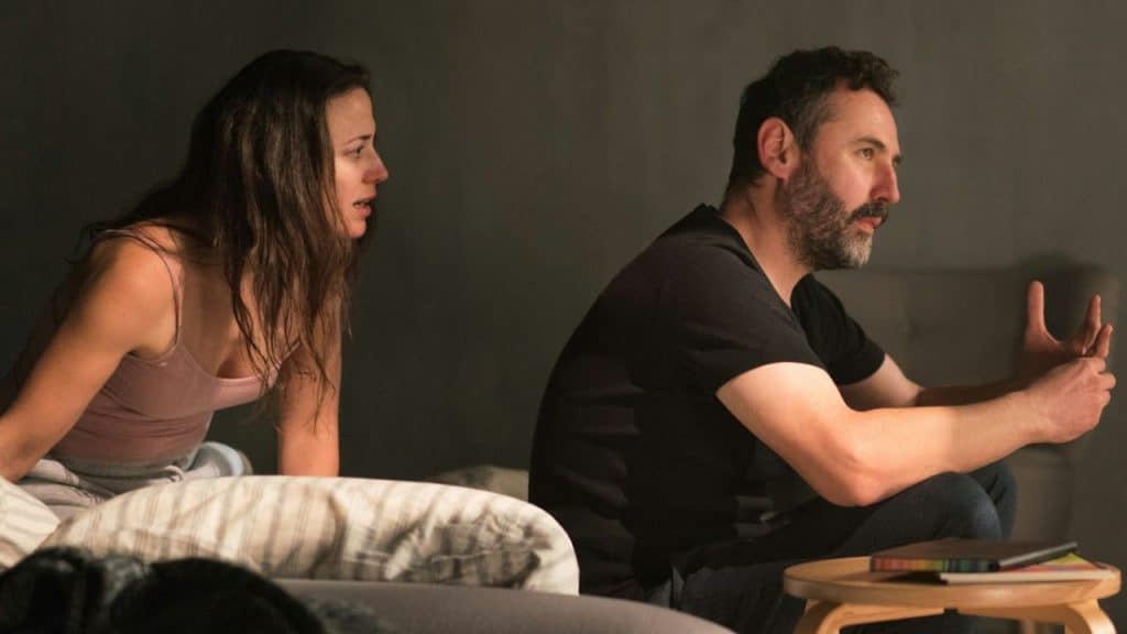 A woman in a tank top and a bearded man sit on a bed or couch, talking intensely.