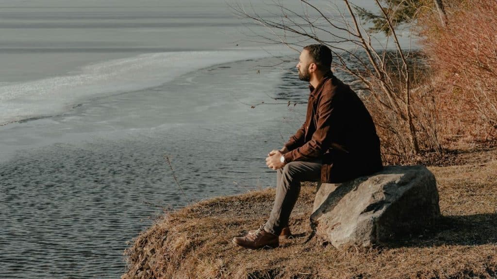 A man in a brown jacket sits on a rock, looking out over a body of water.
