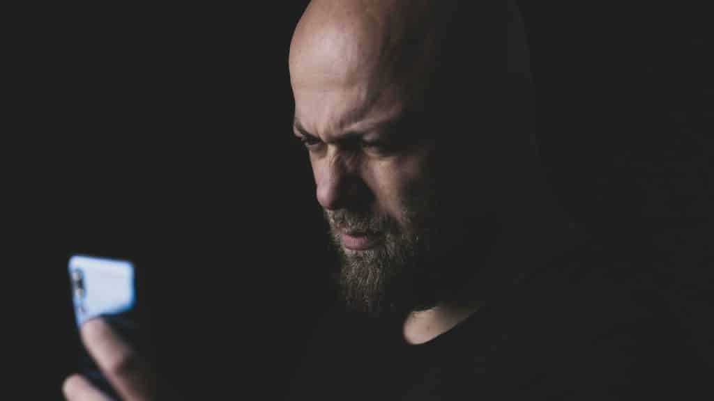 A bald man with a beard looks at a glowing smartphone screen with a furrowed brow.