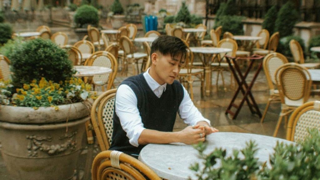 A man sits alone at a round table in an empty outdoor cafe courtyard.