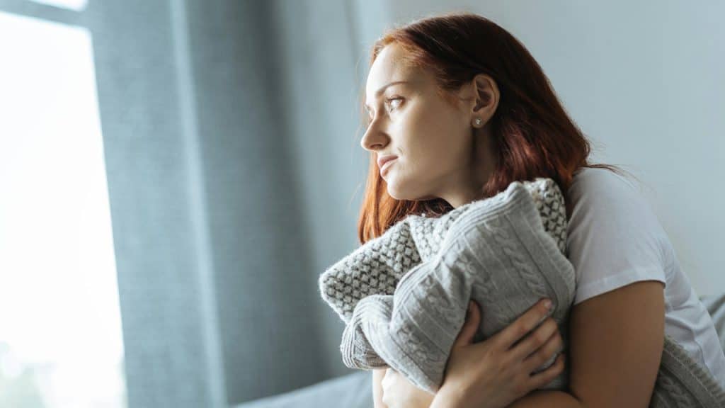 A young woman with red hair sits on a couch, clutching a grey knitted pillow tightly to her chest while gazing thoughtfully out a window.
