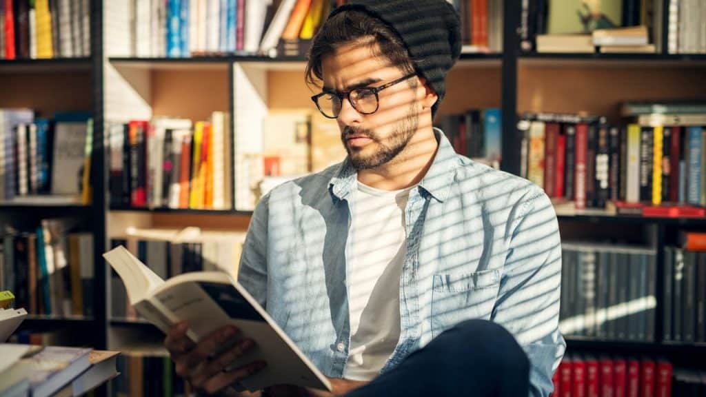 A man wearing glasses and a beanie reading a book in front of a bookshelf.