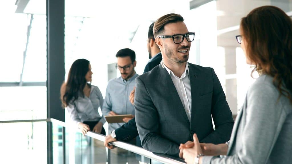 A man in a suit smiles while talking to a woman in an office hallway.