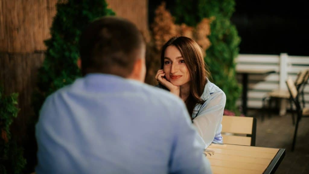 A woman rests her chin on her hand while looking at a man seated outdoors.