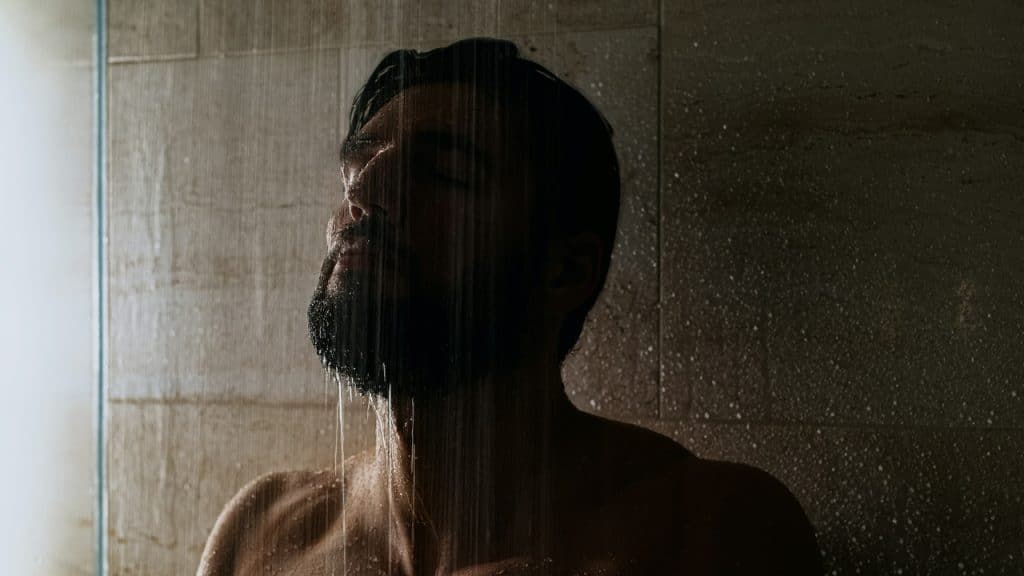 Water falls over the face and shoulders of a bearded man in a dark shower.