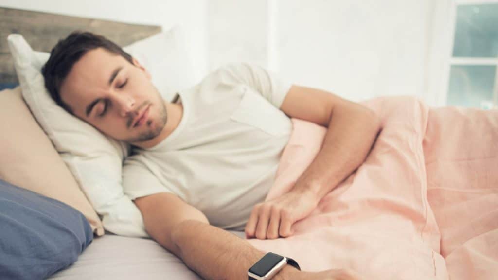 A man sleeping in bed wearing a white t-shirt and a black smartwatch.