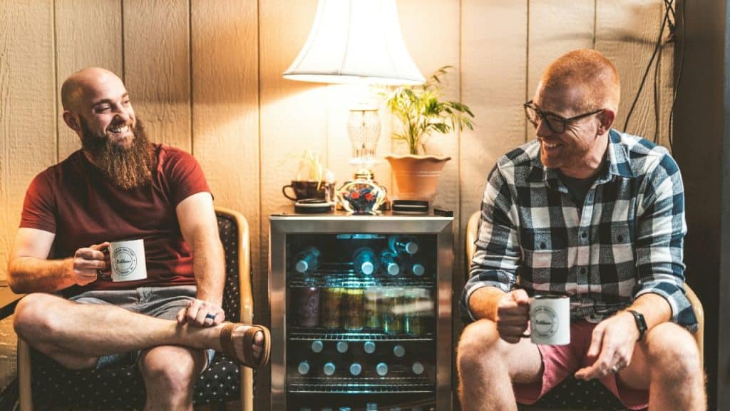 Two men sit in chairs, holding mugs and laughing together in a dimly lit room.