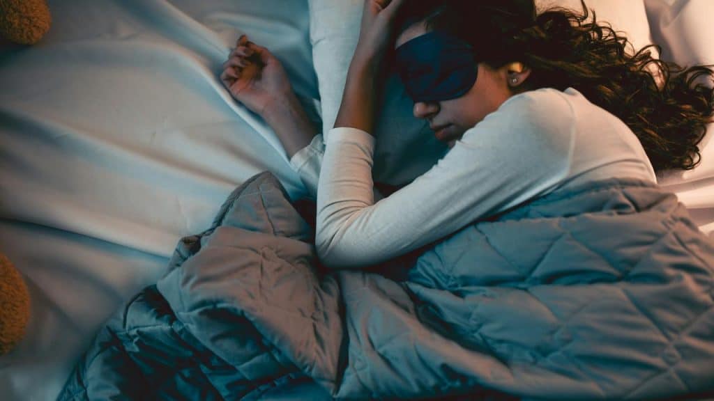 A woman sleeping on her side in bed, wearing an eye mask under a blanket.