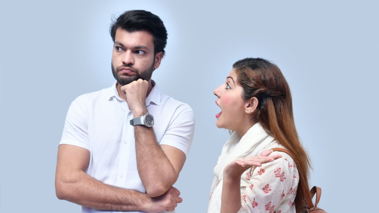 A man with crossed arms looks away while a woman speaks with an open-mouthed expression.