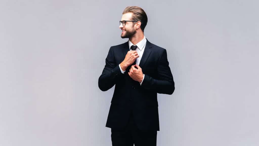 A handsome young man in a full suit adjusting his necktie and smiling.