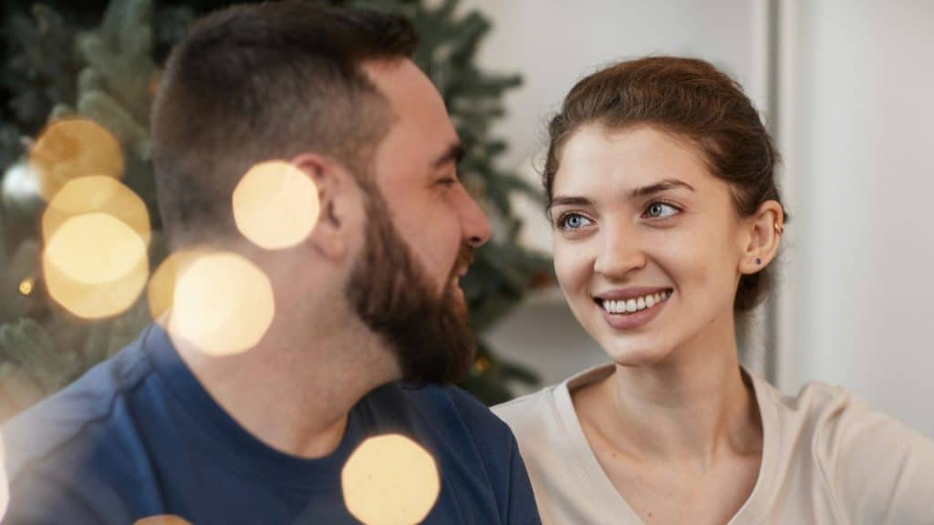 A man and woman look at each other and smile with blurred lights in the foreground.