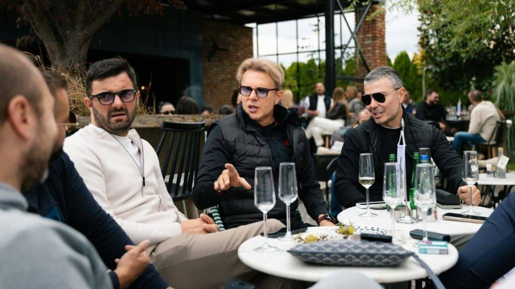 Four men sit at an outdoor restaurant table with drinks, engaged in an intense conversation.