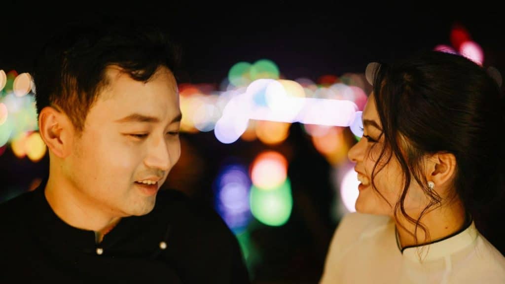 A smiling woman looks toward a man against a background of blurred city lights.