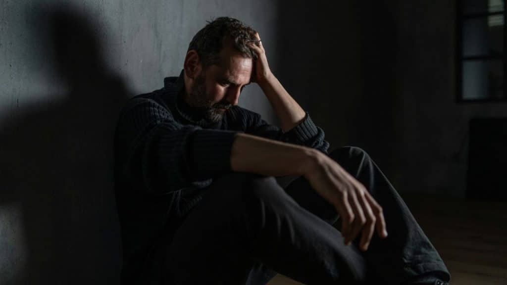 A man sits on the floor leaning against a dark wall with his hand on his head.