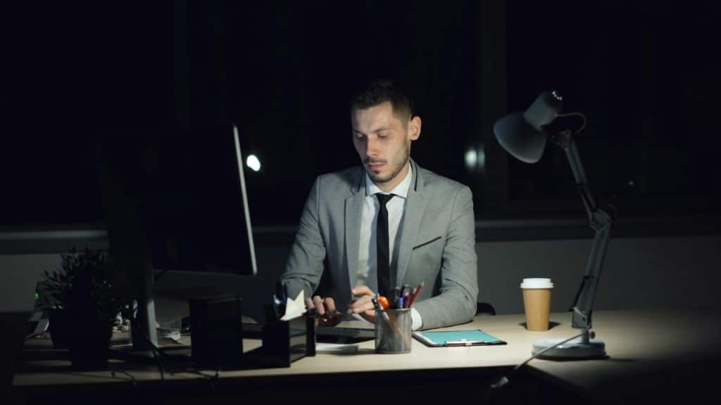 A man in a gray suit sits at a desk working on a computer at night.