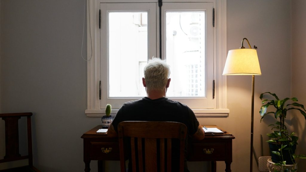 A person with light hair sits at a desk facing a bright window beside a lamp.