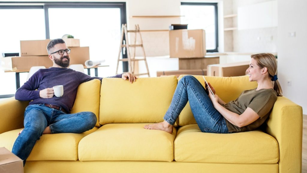 A man and woman sit on a yellow sofa in a room filled with cardboard boxes.
