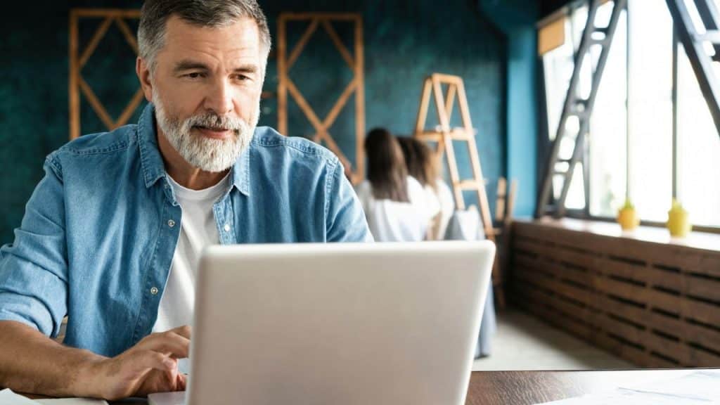 A man with a grey beard sits at a table while using a laptop computer.