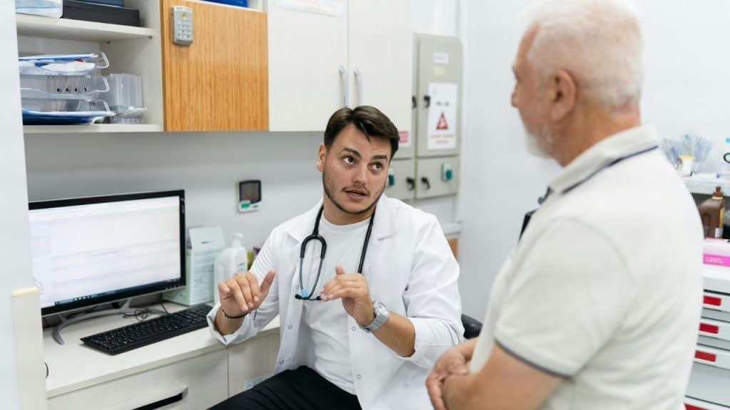 A doctor in a white coat talks to an older patient in a clinical office setting.
