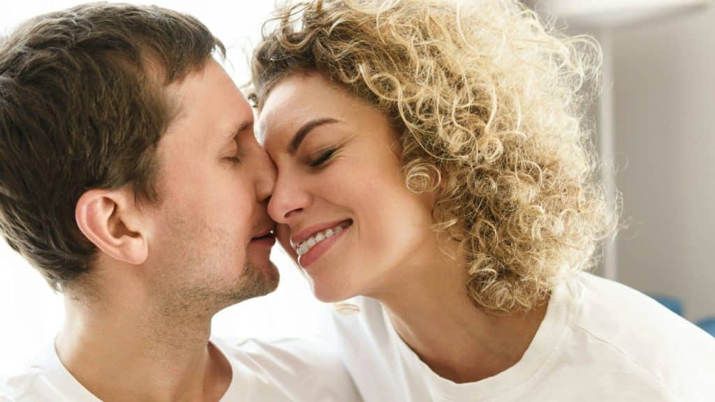 A man and a woman with curly hair touch noses while smiling with eyes closed.