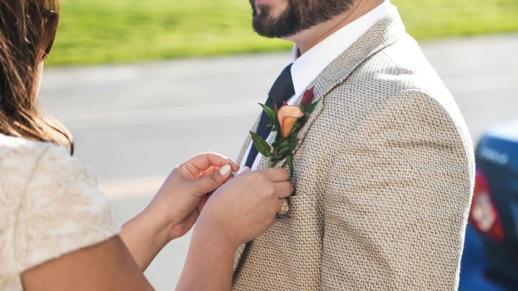 A bride pinning a boutonniere onto a groom’s tan suit jacket.