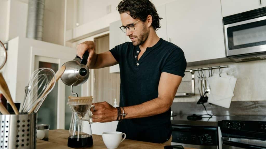 A man with glasses pours hot water into a pour-over coffee maker in a kitchen.