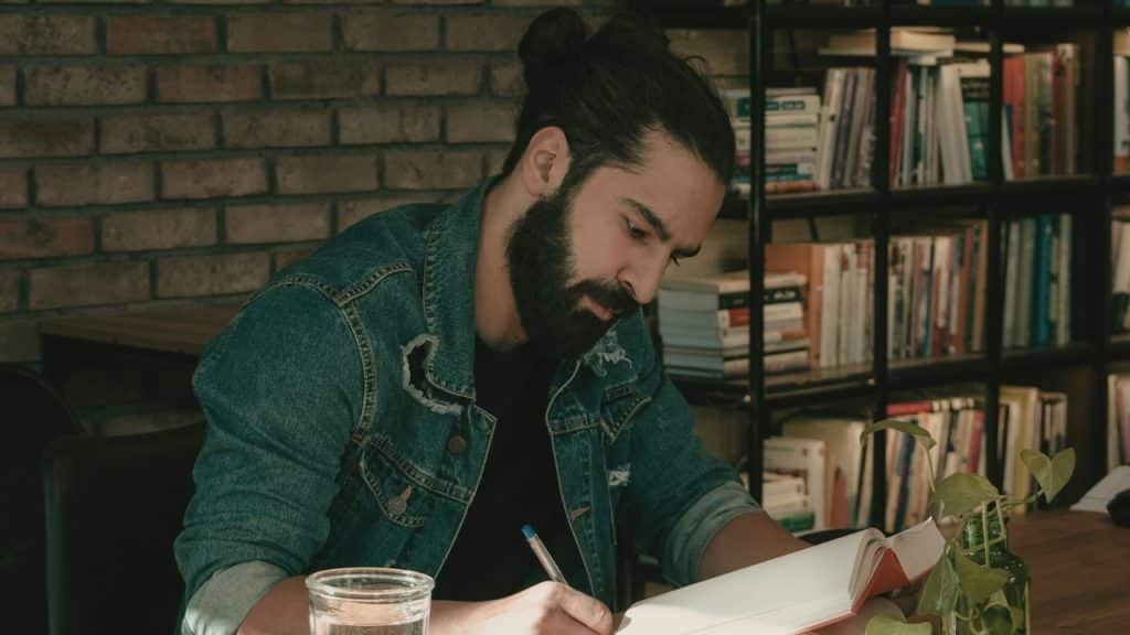 A man with a beard and hair bun writes in a book in a library.