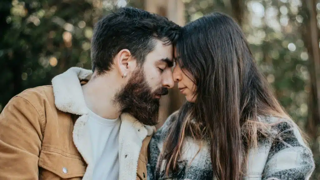 A man and woman sit closely outdoors with their foreheads touching and eyes closed.
