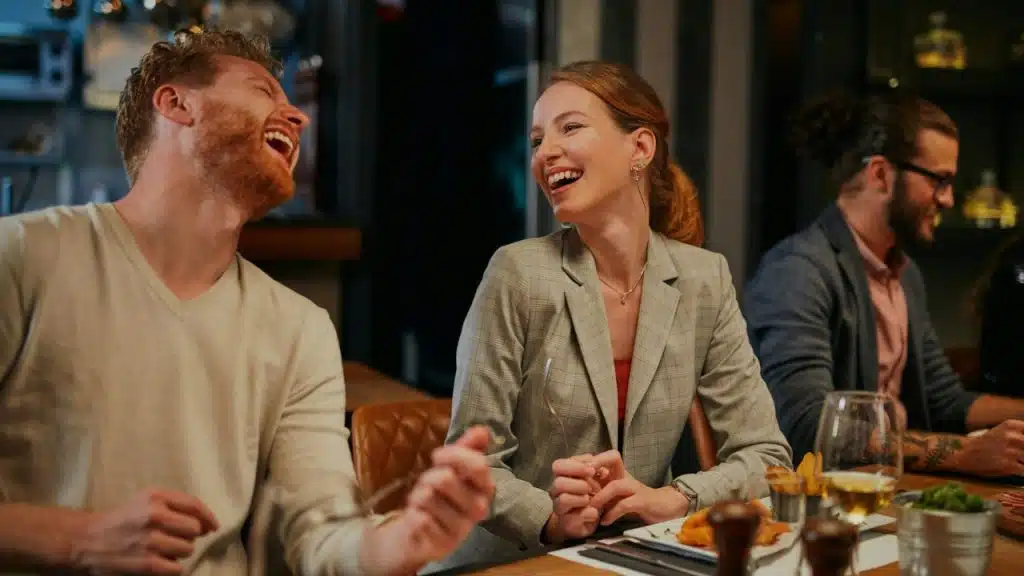A man and woman laugh together while sitting at a restaurant table during a meal.