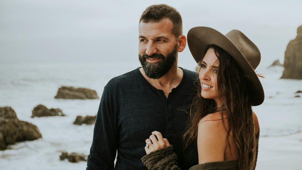 A couple standing close together on a rocky beach, smiling toward the ocean.